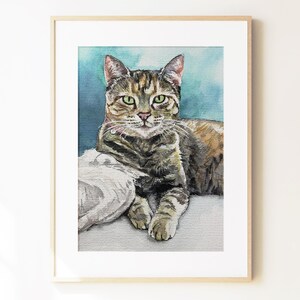 May include: A watercolor painting of a brown tabby cat with green eyes, resting on a white surface against a blue background. The cat is depicted in a realistic style, framed in a light-colored wooden frame with a white mat border.