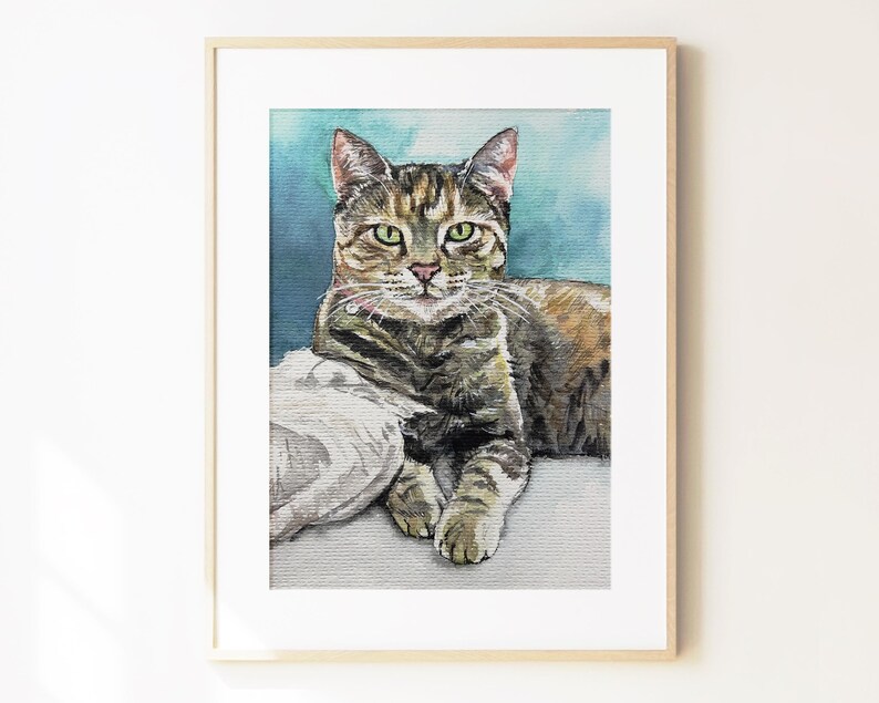 May include: A watercolor painting of a brown tabby cat with green eyes, resting on a white surface against a blue background. The cat is depicted in a realistic style, framed in a light-colored wooden frame with a white mat border.