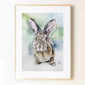 May include: A framed watercolor painting of a brown rabbit with long ears, depicted in a front-facing pose. The artwork features soft green and blue washes in the background and includes a small signature in the bottom right corner of the print.