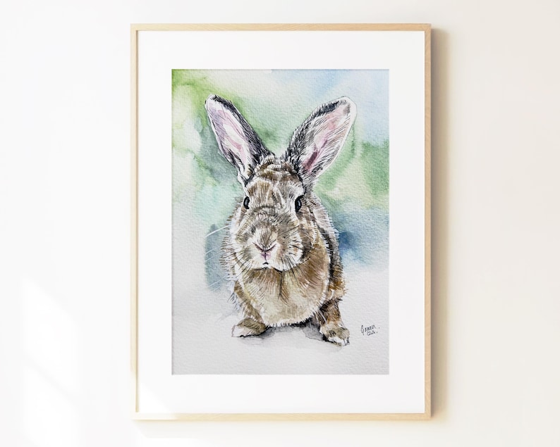 May include: A framed watercolor painting of a brown rabbit with long ears, depicted in a front-facing pose. The artwork features soft green and blue washes in the background and includes a small signature in the bottom right corner of the print.
