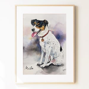 May include: A framed watercolor painting of a white dog with black and tan markings on its head, wearing a red collar with a tag. The dog is sitting with its tongue out. The name Mila is written in black script in the bottom left corner.