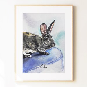 May include: A framed watercolor painting of a dark brown and black rabbit with long ears, positioned on a solid blue surface. The name Ada is written in black cursive script at the bottom of the rabbit's blue perch within the artwork.