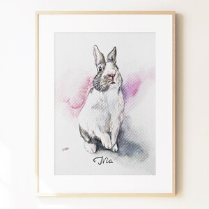 May include: A framed watercolor illustration of a white and grey rabbit with long ears, sitting upright. The rabbit is positioned against a soft pink and grey wash background. The name Nia is written in black cursive script below the rabbit.