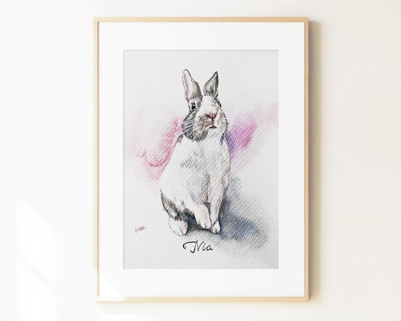 May include: A framed watercolor illustration of a white and grey rabbit with long ears, sitting upright. The rabbit is positioned against a soft pink and grey wash background. The name Nia is written in black cursive script below the rabbit.