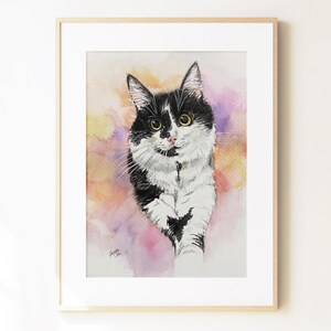 May include: A framed watercolor painting of a black and white cat with yellow eyes. The cat is depicted against a soft, blended background of yellow, orange, and purple watercolor washes. The artwork is enclosed in a light-colored wooden frame.