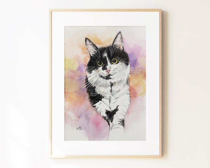 May include: A framed watercolor painting of a black and white cat with yellow eyes. The cat is depicted against a soft, blended background of yellow, orange, and purple watercolor washes. The artwork is enclosed in a light-colored wooden frame.