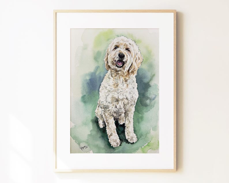 May include: A framed watercolor painting of a cream-colored Goldendoodle dog with curly fur, a black nose, and an open mouth. The dog is centered against a soft, blended background of green and blue watercolor washes, signed in the bottom corner.