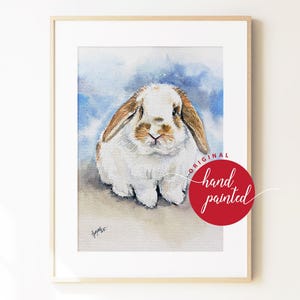 May include: A framed watercolor painting of a white and brown lop-eared rabbit against a blue wash background. A red circular stamp on the lower right reads ORIGINAL hand painted. The artwork is enclosed in a simple light wood frame.
