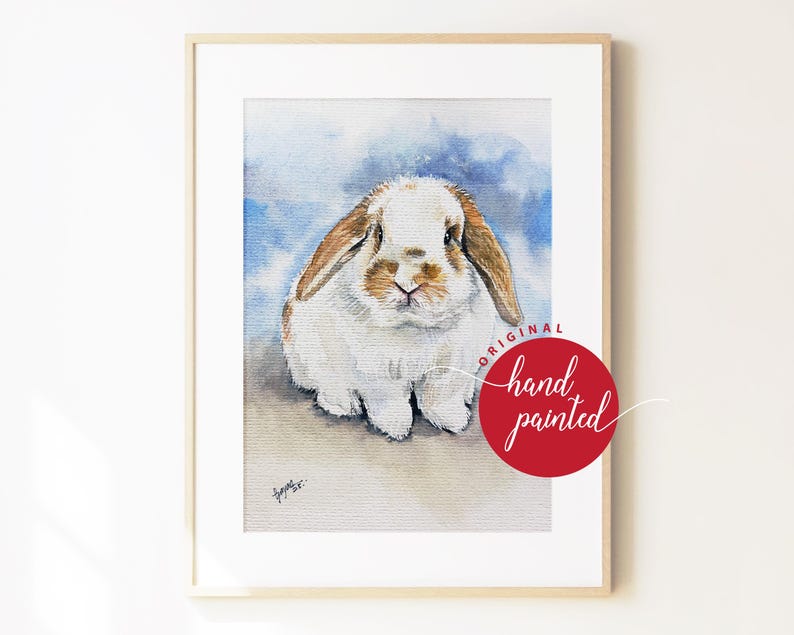 May include: A framed watercolor painting of a white and brown lop-eared rabbit against a blue wash background. A red circular stamp on the lower right reads ORIGINAL hand painted. The artwork is enclosed in a simple light wood frame.
