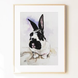 May include: A framed watercolor painting of a black and white rabbit with upright ears. The rabbit has a black nose and eye patches, set against a soft, light purple and white wash background. The artwork is enclosed in a light wood frame.