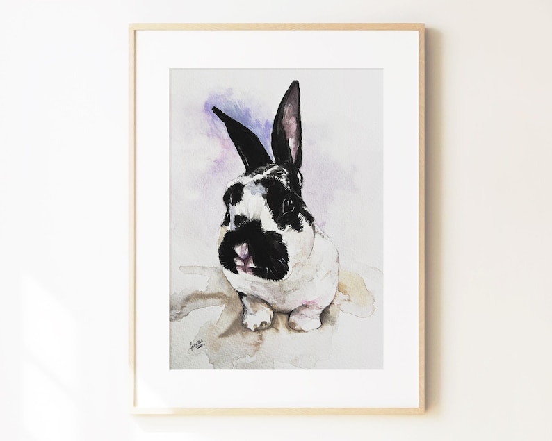 May include: A framed watercolor painting of a black and white rabbit with upright ears. The rabbit has a black nose and eye patches, set against a soft, light purple and white wash background. The artwork is enclosed in a light wood frame.