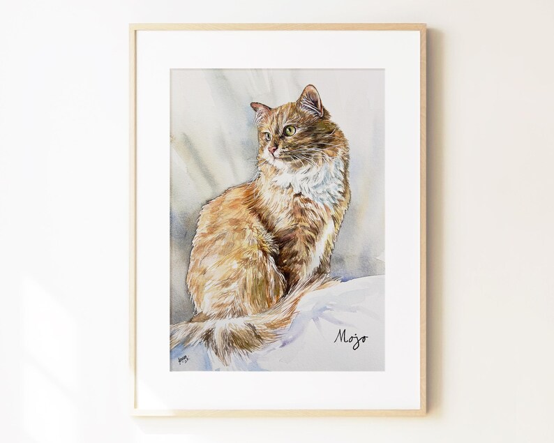 May include: A framed watercolor painting of a long-haired ginger cat with a white chest and green eyes, sitting in a profile pose. The artwork features a soft, neutral-toned background and includes the handwritten name Mojo in the lower right corner.