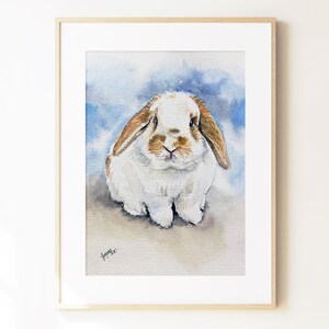 May include: A framed watercolor painting of a lop-eared rabbit with white and light brown fur. The rabbit is centered against a soft, blended blue and white background. A small, dark signature is visible in the bottom left corner of the artwork.