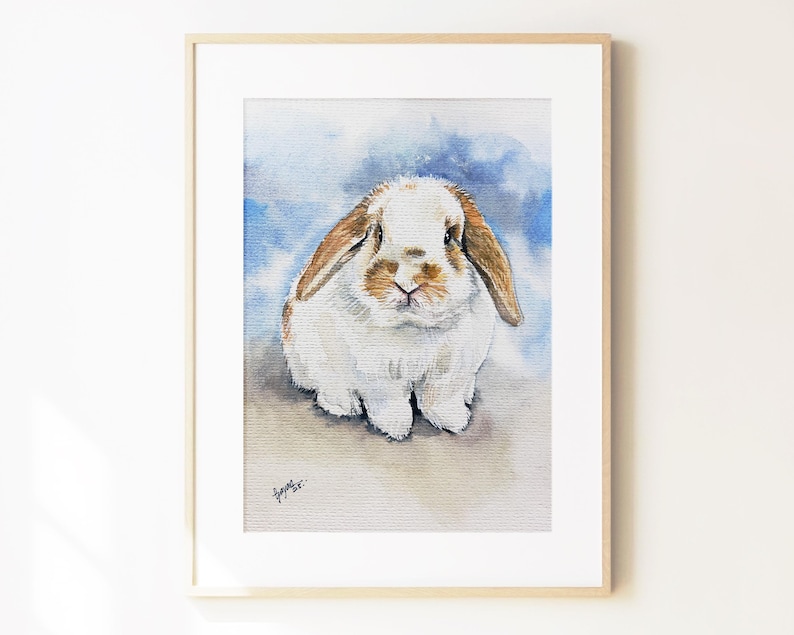 May include: A framed watercolor painting of a lop-eared rabbit with white and light brown fur. The rabbit is centered against a soft, blended blue and white background. A small, dark signature is visible in the bottom left corner of the artwork.