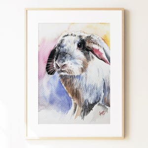 May include: A watercolor painting of a lop-eared rabbit with white, brown, and black fur, set against a soft, abstract background of pink, yellow, and blue washes. The artwork is displayed in a light-colored wooden frame with a white mat border.