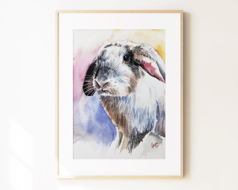 May include: A watercolor painting of a lop-eared rabbit with white, brown, and black fur, set against a soft, abstract background of pink, yellow, and blue washes. The artwork is displayed in a light-colored wooden frame with a white mat border.