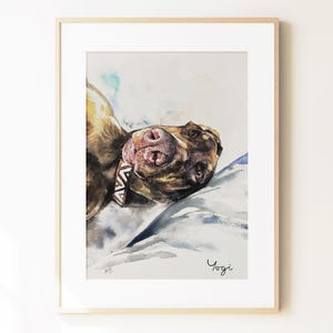 May include: A framed watercolor painting of a brown dog lying down, viewed from a close-up angle. The dog wears a patterned collar and rests on a light-colored surface with blue shadows. The name Yogi is written in black script at the bottom right.