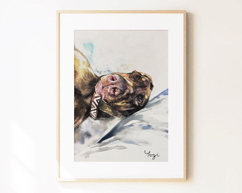 May include: A framed watercolor painting of a brown dog lying down, viewed from a close-up angle. The dog wears a patterned collar and rests on a light-colored surface with blue shadows. The name Yogi is written in black script at the bottom right.