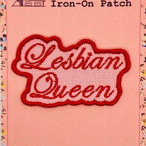 May include: A red embroidered iron-on patch with the words "Lesbian Queen" in a cursive font. The patch is outlined in red and set against a pink background. The text "Pretty In Pulp" and "Iron-On Patch" are also visible.
