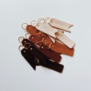May include: A collection of leather keychains with different colors and phrases, including "Cheers", "Play", "Love You", "XOXO", and "The Little Things".