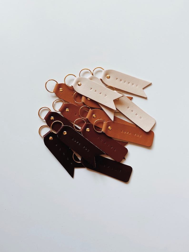 May include: A collection of leather keychains with different colors and phrases, including "Cheers", "Play", "Love You", "XOXO", and "The Little Things".