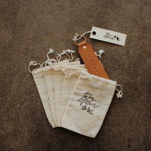 May include: A set of six small, white cotton drawstring bags with the words "twin sparrow" and two birds printed on one bag. A brown leather keychain with the word "CREATE" embossed on it is included.