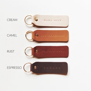 May include: Four leather keychains in different colors: cream, camel, rust, and espresso. Each keychain has a gold ring and a different text embossed on the leather. The text on the keychains reads: "Mama Bear", "San Francisco", "Welcome Home", and "Celebrate".