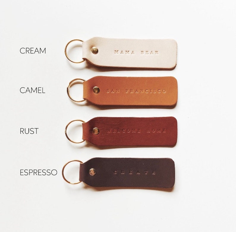 May include: Four leather keychains in different colors: cream, camel, rust, and espresso. Each keychain has a gold ring and a different text embossed on the leather. The text on the keychains reads: "Mama Bear", "San Francisco", "Welcome Home", and "Celebrate".