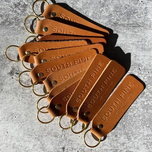 May include: A stack of eight brown leather keychains with gold key rings. The keychains are embossed with the text 'SOUTH PINE' and the number '0175'.