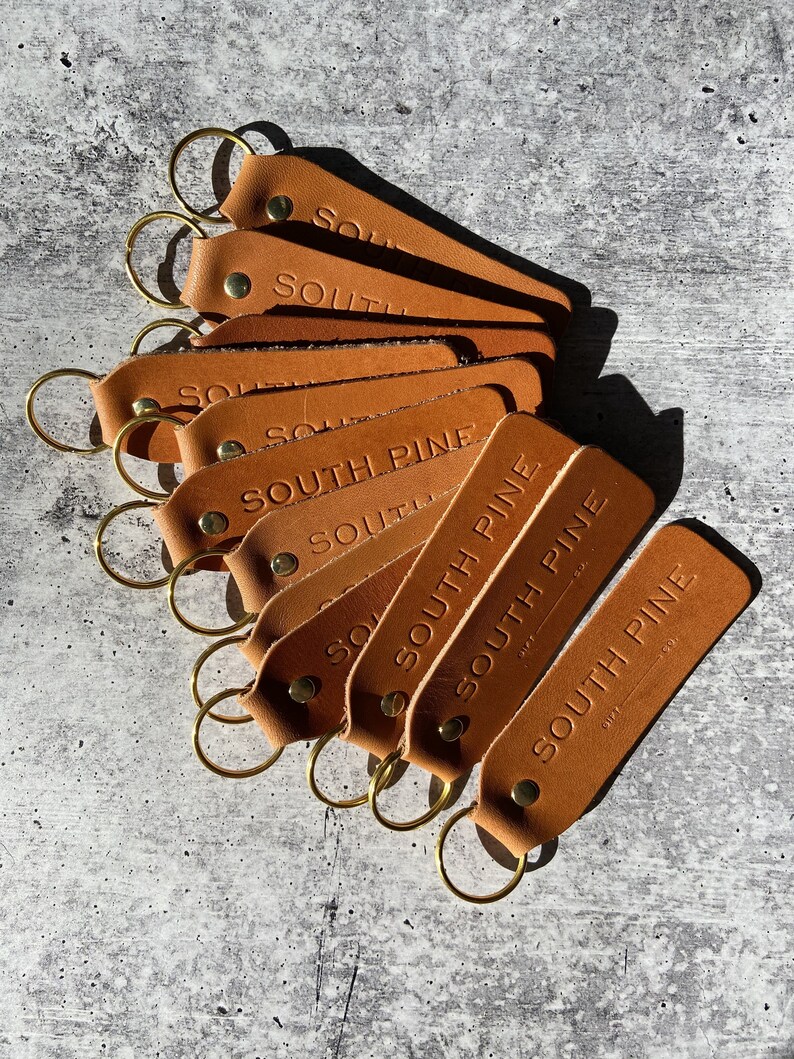 May include: A stack of eight brown leather keychains with gold key rings. The keychains are embossed with the text 'SOUTH PINE' and the number '0175'.