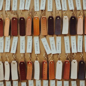 May include: A display of leather keychains with gold rings and white paper tags with handwritten names. The keychains are in various shades of brown, tan, and cream.