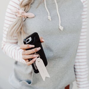 May include: A gray and white striped hoodie with a pink stripe. The hoodie has a white drawstring and a small brown patch on the bottom. A person is holding a black phone case with a key chain that says "Be Kind".