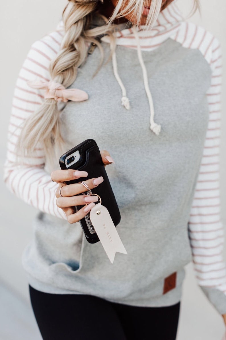 May include: A gray and white striped hoodie with a pink stripe. The hoodie has a white drawstring and a small brown patch on the bottom. A person is holding a black phone case with a key chain that says "Be Kind".