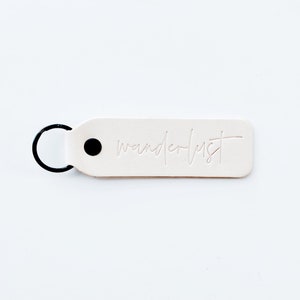 Peut inclure: Porte-clés en cuir blanc avec un anneau en métal noir et un point noir. Le mot "wanderlust" est gravé sur le porte-clés.