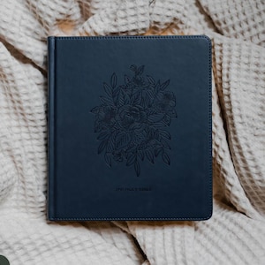 May include: A navy blue hardcover book with a floral design embossed on the cover. The text "THE HOLY BIBLE" is printed at the bottom. The book is lying on a beige, textured surface.