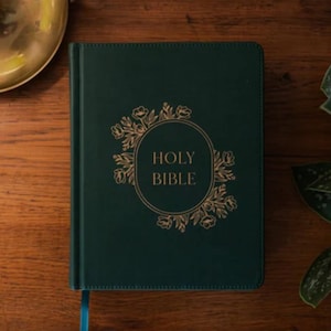 May include: A dark green hardcover Bible with gold lettering and floral accents. The words "HOLY BIBLE" are centered within a gold circular frame. A blue ribbon bookmark is visible. The book is on a wooden surface.