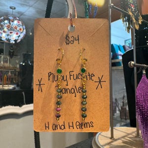 Peut inclure: Boucles d'oreilles pendantes Ruby Fucsite dorées avec des perles vertes et roses. Les boucles d'oreilles sont présentées sur une étiquette marron avec le texte "Ruby Fucsite Dangles" et "Hand and H Gems".