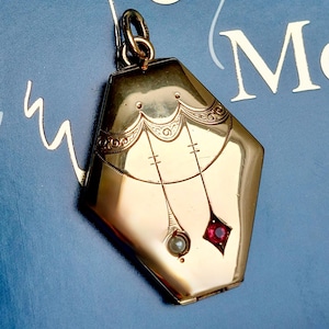 May include: A gold-colored, geometric locket with a pearl and a red gemstone, hanging on a chain. The locket is set against a blue background with the words "Toi & Moi" and dove illustrations in white.
