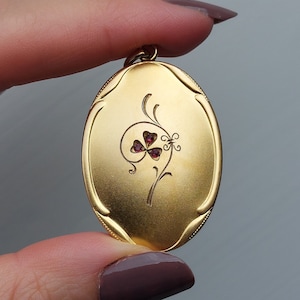 Puede incluir: Un medallón ovalado de oro con un delicado diseño floral y un trébol rojo en el centro.