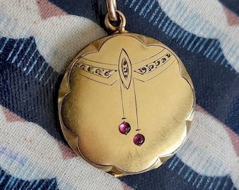 May include: A gold locket necklace with a delicate chain. The locket is round with a scalloped edge and features an intricate design of a dragonfly with two ruby-red stones hanging from its wings.