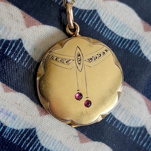 Può includere: Una collana con un ciondolo d'oro e una catena delicata. Il ciondolo è rotondo con un bordo a festoni e presenta un design intricato di una libellula con due pietre rosso rubino che pendono dalle sue ali.