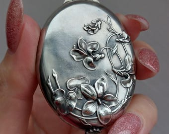 May include: Oval-shaped silver pendant with a floral design. The pendant features embossed flowers and leaves, with a loop at the top for a chain. The metal has a polished, reflective surface, and the floral details are intricately carved.