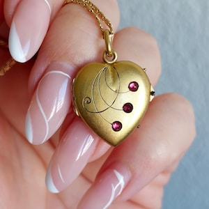May include: A gold heart-shaped locket pendant with three red gemstones. The locket has an ornate design and is suspended from a delicate gold chain. The locket is held by a person with manicured nails.