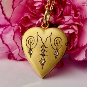 May include: A gold-toned heart-shaped locket pendant suspended from a matching gold-toned chain. The front surface of the locket features an engraved symmetrical pattern with three small, clear, inset stones arranged in a vertical and diagonal design.