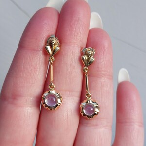 May include: A pair of gold drop earrings featuring a small, heart-shaped top element connected to a slender bar that leads to a circular base. Each base is set with a smooth, round, pale pink cabochon stone surrounded by a scalloped gold edge.
