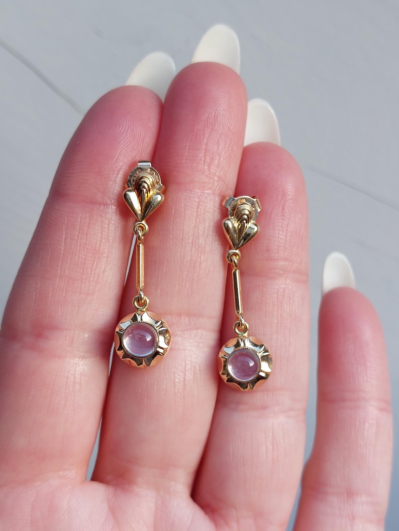 May include: A pair of gold drop earrings featuring a small, heart-shaped top element connected to a slender bar that leads to a circular base. Each base is set with a smooth, round, pale pink cabochon stone surrounded by a scalloped gold edge.