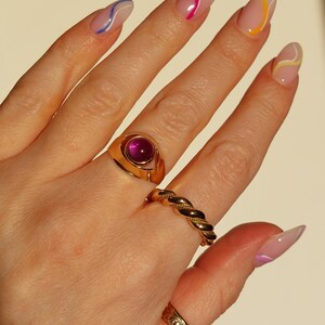 May include: A hand wearing several gold rings, including a signet ring with a large, round, pink gemstone. Other rings include a twisted band and a patterned band. The nails are painted with a clear base and colorful wavy lines.