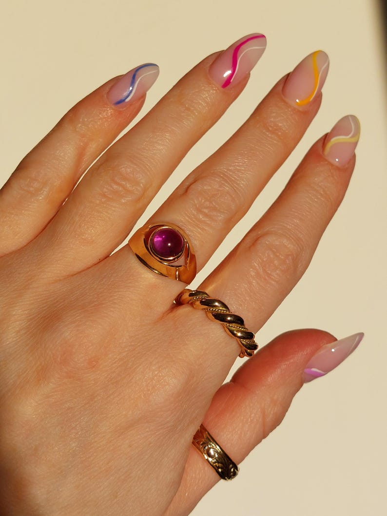 May include: A hand wearing several gold rings, including a signet ring with a large, round, pink gemstone. Other rings include a twisted band and a patterned band. The nails are painted with a clear base and colorful wavy lines.