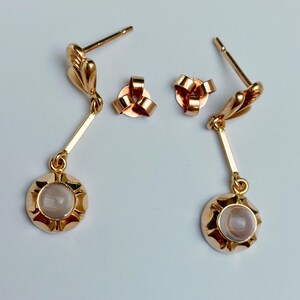 May include: A pair of gold-toned drop earrings featuring a post back and a small, round, light-colored cabochon stone set within a scalloped metal frame. Each earring hangs from a thin, straight metal bar attached to a decorative leaf-shaped top.