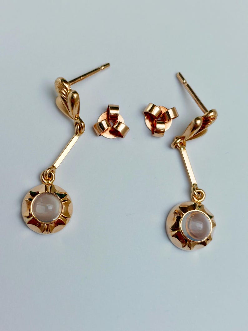 May include: A pair of gold-toned drop earrings featuring a post back and a small, round, light-colored cabochon stone set within a scalloped metal frame. Each earring hangs from a thin, straight metal bar attached to a decorative leaf-shaped top.